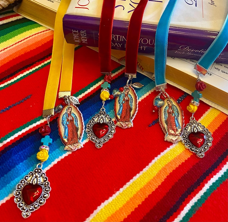 May include: Four colorful ribbon bookmarks with silver charms featuring an image of the Virgin Mary, a red heart, and various beads. The bookmarks are displayed on a colorful striped fabric.