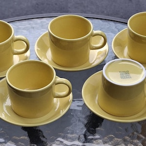 May include: A set of five yellow ceramic cups and saucers. Four cups are upright, and one cup is inverted to show the base with the printed text "KILTA FINLAND TEEMA". Each cup features a simple cylindrical body with a side handle and a matching saucer.