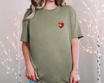 Könnte beinhalten: Olivgrünes T-Shirt mit einem kleinen gestickten Design eines roten Herzens mit Flammen und einer weißen Blume. Das kurzärmelige Shirt ist aus einem weichen Material gefertigt und hat eine lockere Passform.