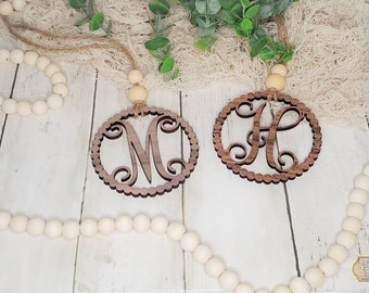 May include: Two wooden monogram ornaments, one with the letter "M" and the other with the letter "A". Each ornament has a scalloped edge and is suspended by a jute string with a wooden bead. Decorative wooden beads and greenery are in the background.