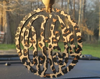 May include: A wooden car charm with a leopard print design and a monogram in the center. The charm is hanging from a string with wooden beads.