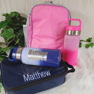 May include: A pink lunch bag with a blue and white water bottle and a blue lunchbox. The lunch bag and lunchbox are personalized with the name "Matthew".
