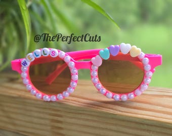 May include: Bright pink sunglasses featuring round frames decorated with a border of small white beads. The left lens is accented with the name WELLS in colorful letter beads, while the right lens features a row of pastel heart-shaped beads.