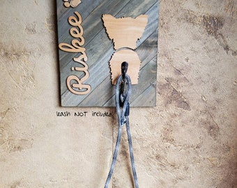 May include: Wooden dog leash holder with a silhouette of a Yorkshire Terrier and the name "Rishkee" written in cursive. The holder is mounted on a wall with a gray and brown wood grain finish. A blue and white rope leash is hanging from the holder. The text "Leash not included" is written below the holder.