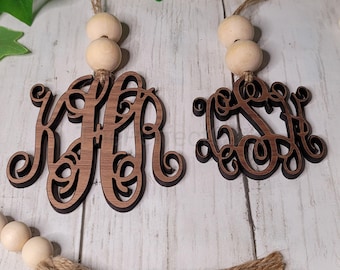 May include: A wooden bead garland with two wooden monogram ornaments, one with the letters "KJR" and the other with the letters "LS". The garland has a brown jute tassel.