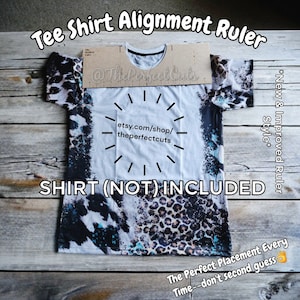 May include: A wooden tee shirt alignment ruler with a white tee shirt with a black and white animal print design on top of it. The ruler has the text "The Perfect Cuts" and "etsy.com/shop/theperfectcuts" printed on it. The text "*New & Improved Ruler Style*" is printed on the right side of the image. The text "SHIRT (NOT) INCLUDED" is printed below the ruler. The text "The Perfect Placement Every Time--don't second guess" is printed at the bottom of the image.