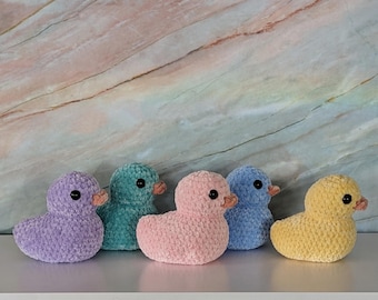 May include: Five crocheted plush duck toys in different colors: lavender, teal, pink, blue, and yellow. The ducks are arranged in a row on a light marble surface.