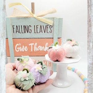 May include: A set of four felt pumpkins in pink, green, white, and lavender with floral accents. The pumpkins are arranged on a white pedestal with a wooden sign that reads "Falling Leaves Give Thanks".