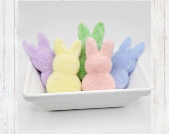 May include: Five felt bunny figurines in a white square dish. The bunnies are in shades of purple, yellow, green, pink, and blue.