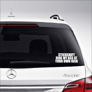 May include: A white car with a black rear window sticker that reads "STICKSHIFT RIDE MY ASS AT YOUR OWN RISK".