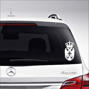 May include: White car decal with a white eagle with a crown on its head and a cross on its chest. The eagle is holding a cross in its talons. The decal is on the back window of a white car. The car has the word "4MATIC" on the back.