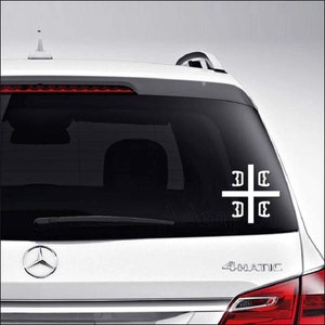 May include: White vinyl decal of a cross with the letters "DC" on each arm. The decal is on the back window of a silver car.