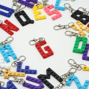 May include: A collection of colorful letter-shaped keychains in various colors, including red, yellow, blue, pink, green, black, and purple. Each keychain features a silver clasp. The letters are designed to resemble building blocks.