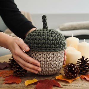 May include: A handmade acorn-shaped basket in olive green and tan. The top is olive green, and the bottom is tan, both with a textured, knitted appearance. The basket is surrounded by fall leaves, pine cones, and candles, creating a seasonal theme.