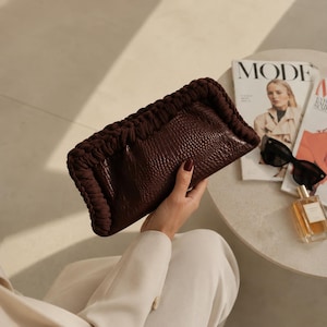 Könnte beinhalten: Eine dunkelbraune Clutch mit strukturierter Lederoberfläche und geflochtenem Rand. Die Tasche wird von einer Person in weißer Kleidung gehalten. Sonnenbrille, Parfüm und eine Zeitschrift liegen auf einem Tisch in der Nähe.