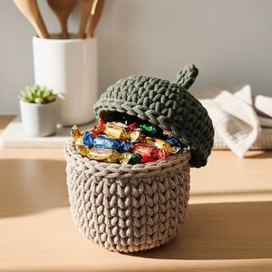 May include: A handmade, beige crochet basket shaped like an acorn, filled with colorful wrapped candies. The basket has a dark green lid, also crocheted, with a stem detail. The basket sits on a wooden surface.
