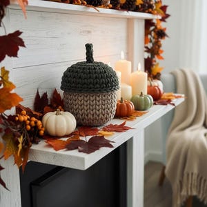May include: A cozy autumn-themed mantel display. A large, two-toned, crocheted acorn sits among small pumpkins, candles, and fall foliage. The color palette includes shades of green, beige, orange, and white, creating a warm and inviting atmosphere.