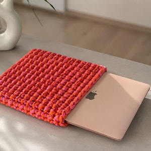 Peut inclure: Une housse pour ordinateur portable rectangulaire fabriquée en fil épais et volumineux, alternant des tons orange vif et rose éclatant. La housse présente un motif en maille crochetée dense et texturée, contenant un ordinateur rose doré.