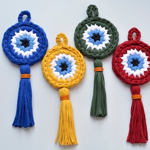 Könnte beinhalten: Vier gehäkelte Glücksbringer mit Quasten. Die Glücksbringer sind blau, gelb, grün und rot. Jeder Glücksbringer hat ein weißes und blaues Auge mit einer schwarzen Pupille.