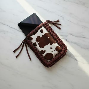 May include: A brown and white cowhide pattern tablet sleeve with a dark brown crocheted border. The sleeve has a rectangular shape and is open at the top, with dark brown ties on each side. A black tablet is partially visible inside.