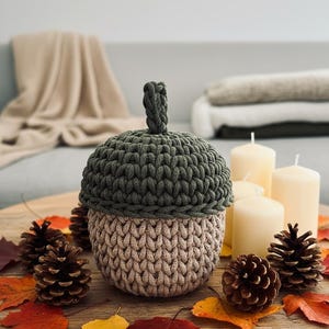 May include: A handmade acorn-shaped container with a dark green top and a tan base, crafted from a thick, textured yarn. The container is surrounded by pine cones, autumn leaves, and candles, creating a fall-themed display.