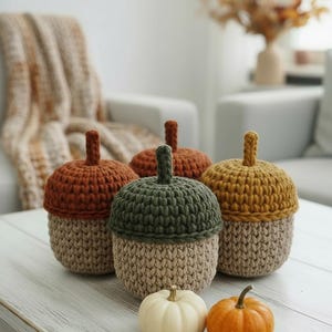 May include: Four crocheted acorn-shaped containers in shades of brown, green, and yellow, with a beige base. Two small pumpkins, one white and one orange, sit in front. The items are arranged on a white wooden table.