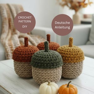 May include: Crocheted acorn-shaped containers in various colors: brown, green, and yellow. The acorn caps are a darker shade than the base. Two small pumpkins sit in front. Text on the image says "CROCHET PATTERN DIY" and "Deutsche Anleitung".