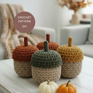 Può includere: Quattro contenitori a forma di ghianda all'uncinetto, nei toni del marrone, verde e giallo, con basi beige, sono su una superficie di legno bianco. Due piccole zucche, una bianca e una arancione, sono davanti. Il testo "CROCHET PATTERN DIY" è incluso.