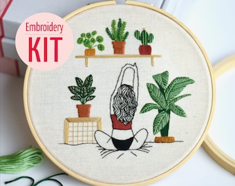 May include: Embroidery kit showing a woman in a yoga pose with embroidered plants. The design is on cream fabric, framed in a wooden hoop. The text "Embroidery KIT" is in a pink circle. The kit includes green thread.