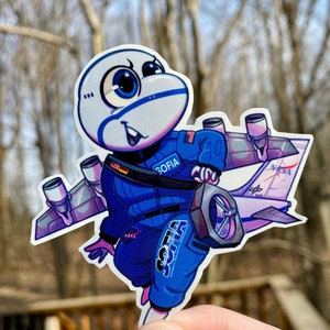 Puede incluir: Una pegatina con un personaje de dibujos animados con un traje de vuelo azul de la NASA, con el nombre "SOFIA" en el traje. El personaje tiene una cabeza y alas con forma de avión, sosteniendo un panel de control. La pegatina tiene un fondo blanco.