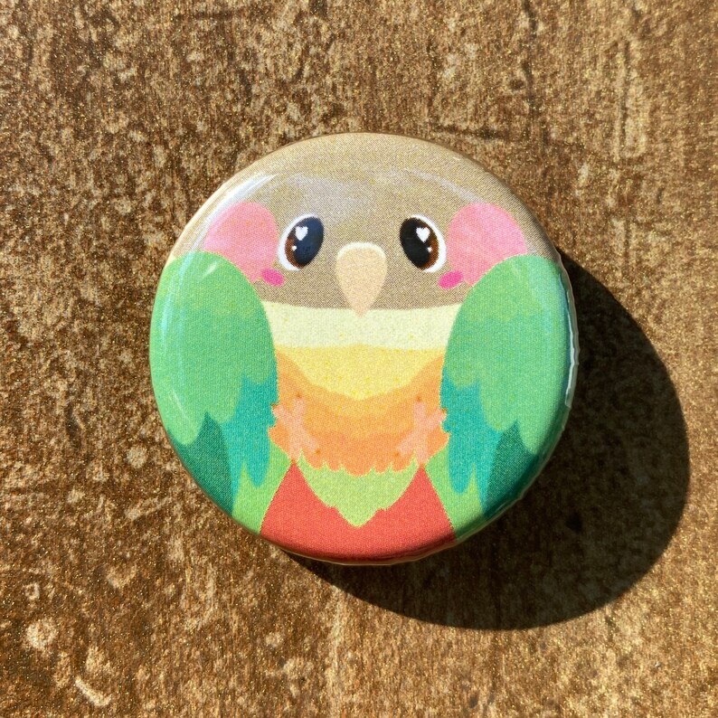 May include: A circular button with a cartoon illustration of a green cheek conure. The bird has a pink face, brown eyes, and a yellow, orange, and red chest. The button has a metallic gold border.
