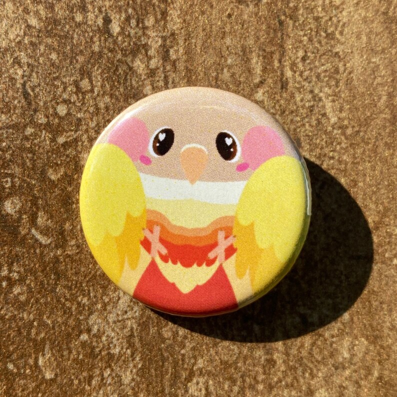 May include: A circular badge featuring a cartoon bird design. The bird has a beige head, pink cheeks, and yellow wings. The bird's chest is a gradient of yellow, orange, and red. The badge is set against a textured brown surface.