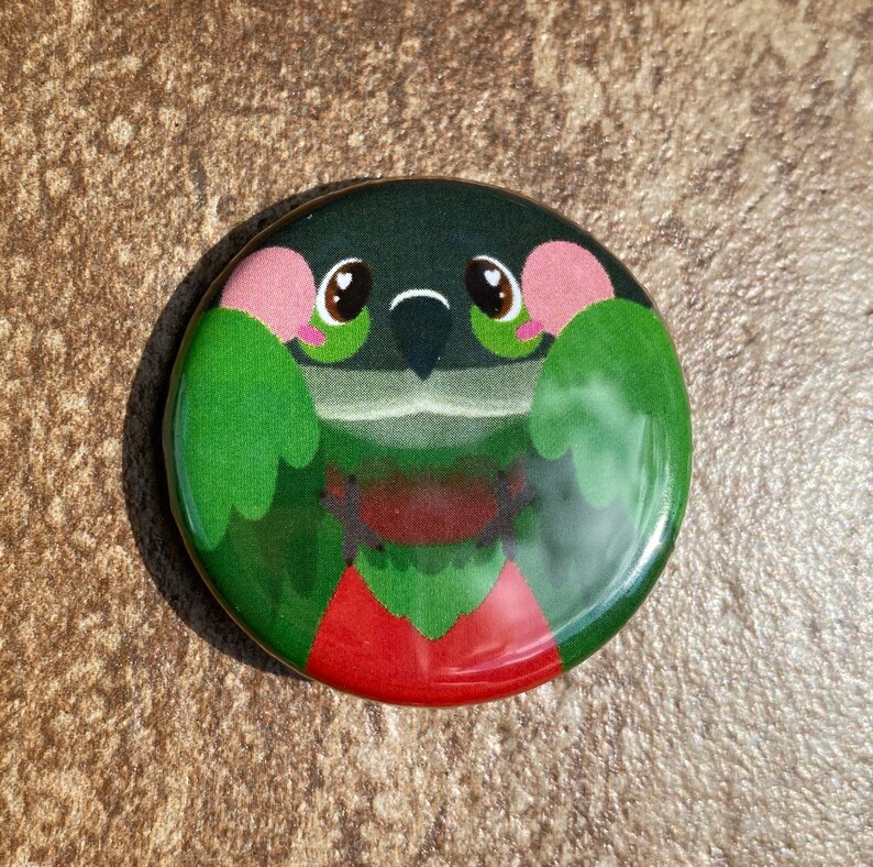 May include: A circular badge with a cartoon bird design. The bird has green feathers, pink cheeks, and a red chest. The badge has a glossy finish and is set against a dark green background. The design is likely printed on metal.