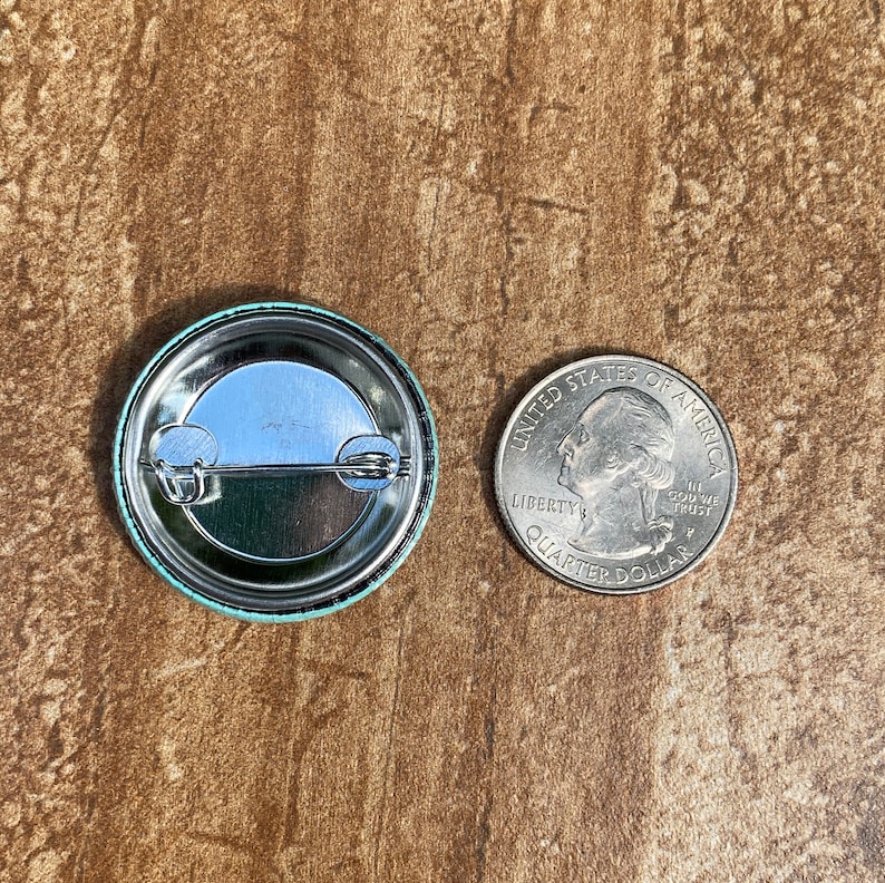 May include: A round button badge with a light blue border and a silver metal back, next to a U.S. quarter coin. The badge's back has a safety pin clasp. The coin is approximately 2.4 cm in diameter.