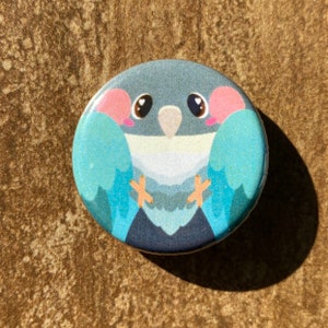 May include: A circular button featuring a cartoon bird illustration. The bird has a blue body, pink cheeks, and large eyes. Its wings are light blue, and its feet are orange. The button is set against a textured, brown background.