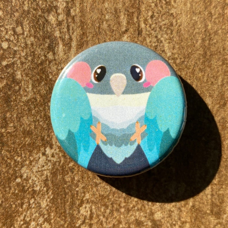 May include: A circular button featuring a cartoon bird illustration. The bird has a blue body, pink cheeks, and large eyes. Its wings are light blue, and its feet are orange. The button is set against a textured, brown background.
