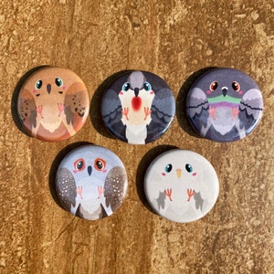 Puede incluir: Cinco botones redondos con ilustraciones de pájaros de dibujos animados. Los botones presentan varios pájaros en tonos marrones, grises y blancos, cada uno con expresiones y detalles únicos. Los botones están dispuestos sobre una superficie marrón texturizada.