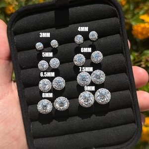 May include: A black velvet jewelry display tray containing eight pairs of round, silver-toned stud earrings with halo settings. The earrings are labeled with sizes 3mm, 4mm, 5mm, 6mm, 6.5mm, 7.5mm, 8mm, and 9mm, indicating their respective diameters.
