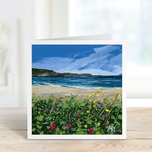 a card sits on a windowsill, the card depicts pink and yellow flowers in the foreground with a beautiful beach and blue sky.