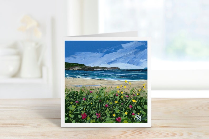 a card sits on a windowsill, the card depicts pink and yellow flowers in the foreground with a beautiful beach and blue sky.