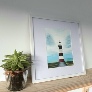 Puede incluir: Pintura enmarcada de un faro con rayas negras, blancas y rojas. La obra de arte está ambientada contra un cielo azul con nubes y un mar turquesa. Una planta suculenta verde en una maceta de vidrio está a la izquierda.