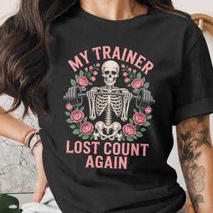 May include: Black t-shirt with a graphic of a skeleton lifting weights, encircled by pink roses. The text reads "MY TRAINER LOST COUNT AGAIN" in pink. A distinctive design for gym-goers.