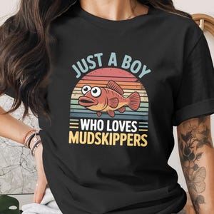 May include: Black t-shirt with a colorful graphic featuring a cartoon mudskipper fish and the text "JUST A BOY WHO LOVES MUDSKIPPERS". The design includes a sunset background with blue, orange, and yellow hues.