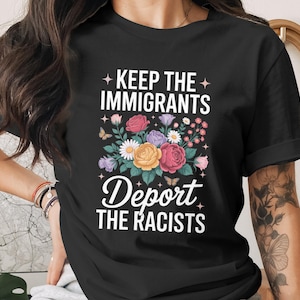 May include: Black t-shirt with the text "KEEP THE IMMIGRANTS, DEPORT THE RACISTS" in white. The shirt features a floral design with roses, daisies, and butterflies in various colors.