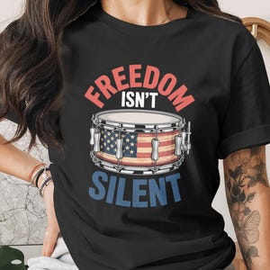 Könnte beinhalten: Schwarzes T-Shirt mit dem Text "FREEDOM ISN'T SILENT" in Rot und Blau. Ein Schlagzeug mit amerikanischem Flaggen-Design ist abgebildet. Das T-Shirt hat einen Rundhalsausschnitt und kurze Ärmel.