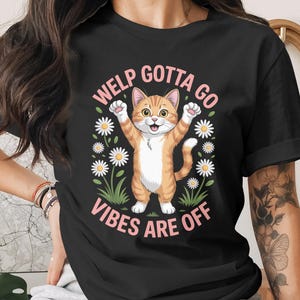 Puede incluir: Camiseta negra con un diseño de gato atigrado naranja de dibujos animados con los brazos levantados. El texto "WELP GOTTA GO VIBES ARE OFF" está en rosa, rodeado de margaritas.