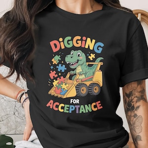 May include: Black t-shirt featuring a cartoon dinosaur driving a yellow front-end loader. The dinosaur is smiling and surrounded by colorful puzzle pieces. Text reads "DIGGING FOR ACCEPTANCE" in rainbow colors.