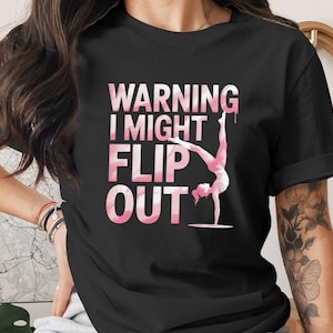 Puede incluir: Camiseta negra con el texto "WARNING I MIGHT FLIP OUT" en rosa y blanco. El gráfico presenta una gimnasta haciendo el pino. La camiseta es una prenda informal.