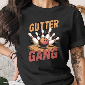 May include: Black t-shirt featuring a bowling design. The graphic includes a bowling ball, pins, and the words "Gutter Gang" in orange and red. The design is set against a wooden floor background.