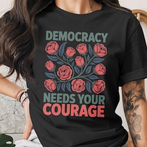 May include: Black t-shirt with the text "DEMOCRACY NEEDS YOUR COURAGE" in green and red. The shirt features a floral design with red roses and blue leaves, creating a bold and political statement.